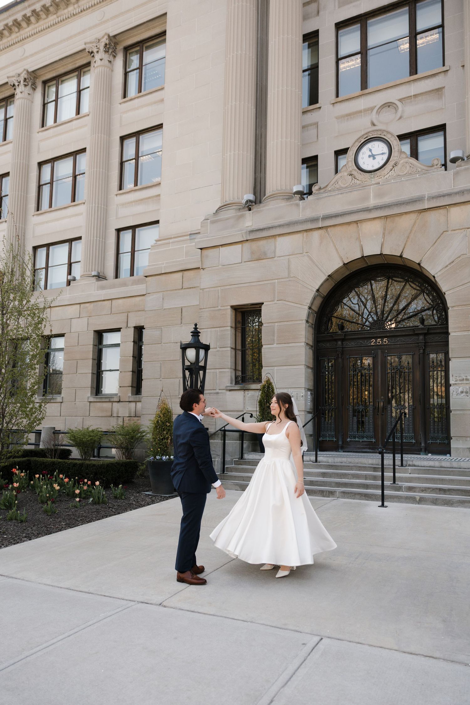 Bride and groom crossing a city street crosswalk in wedding attire, holding hands near a traffic light and trees.