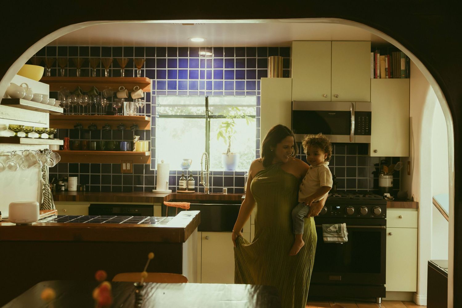 A woman holding a child in a cozy kitchen with blue tile backsplash, wooden shelves, and arched doorway.