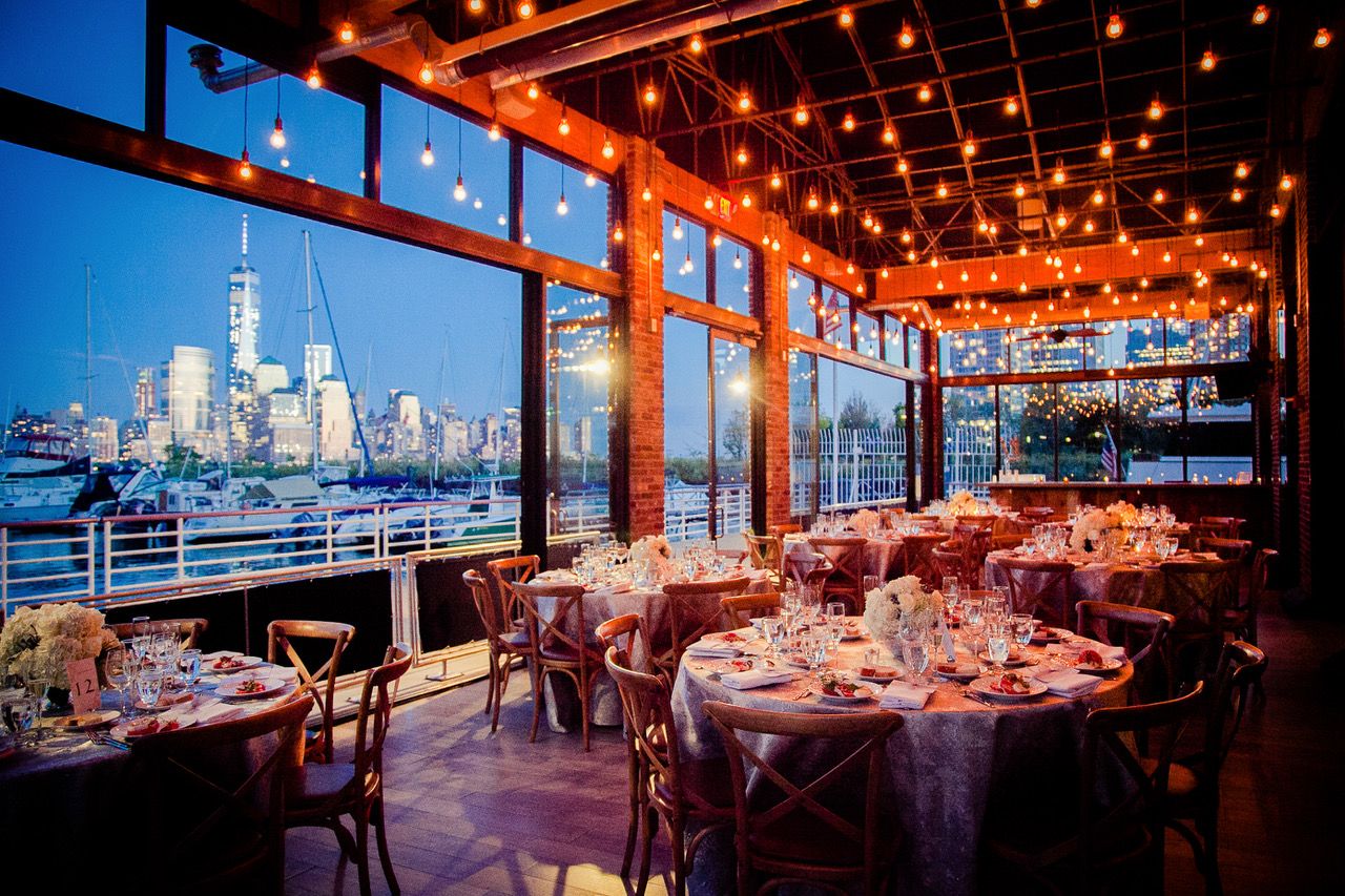 An elegant waterfront restaurant event space with string lights and city skyline views set for a formal dinner reception.
