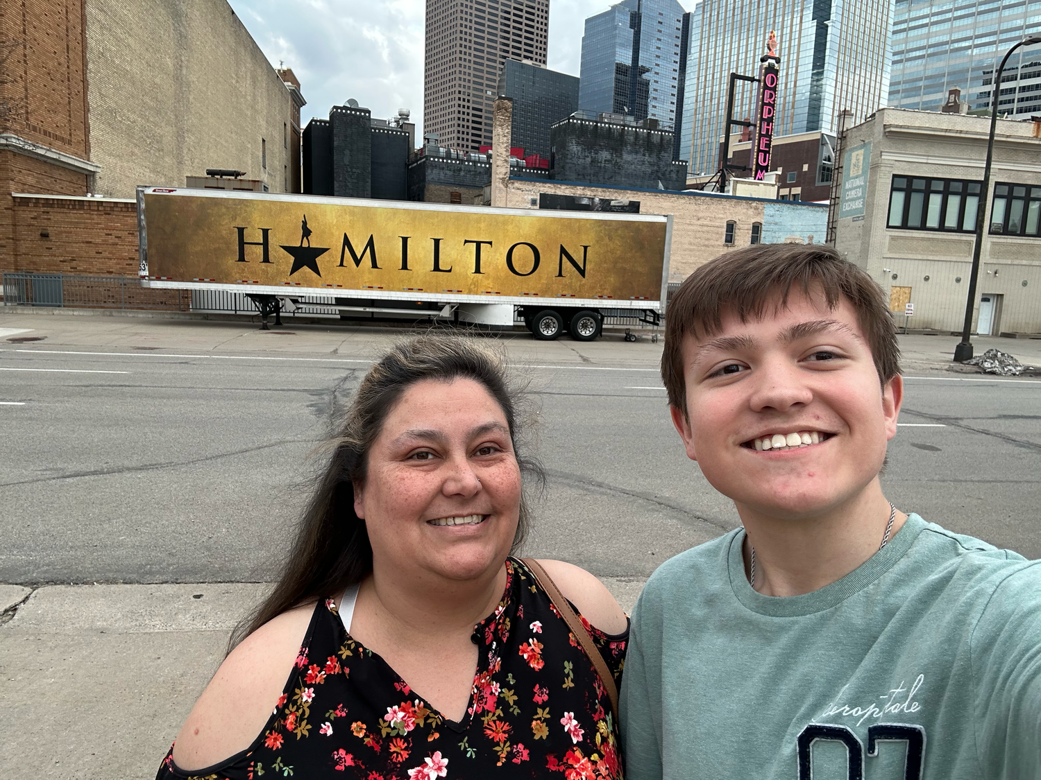 Two people smiling in front of a Hamilton musical touring production truck in a city setting.