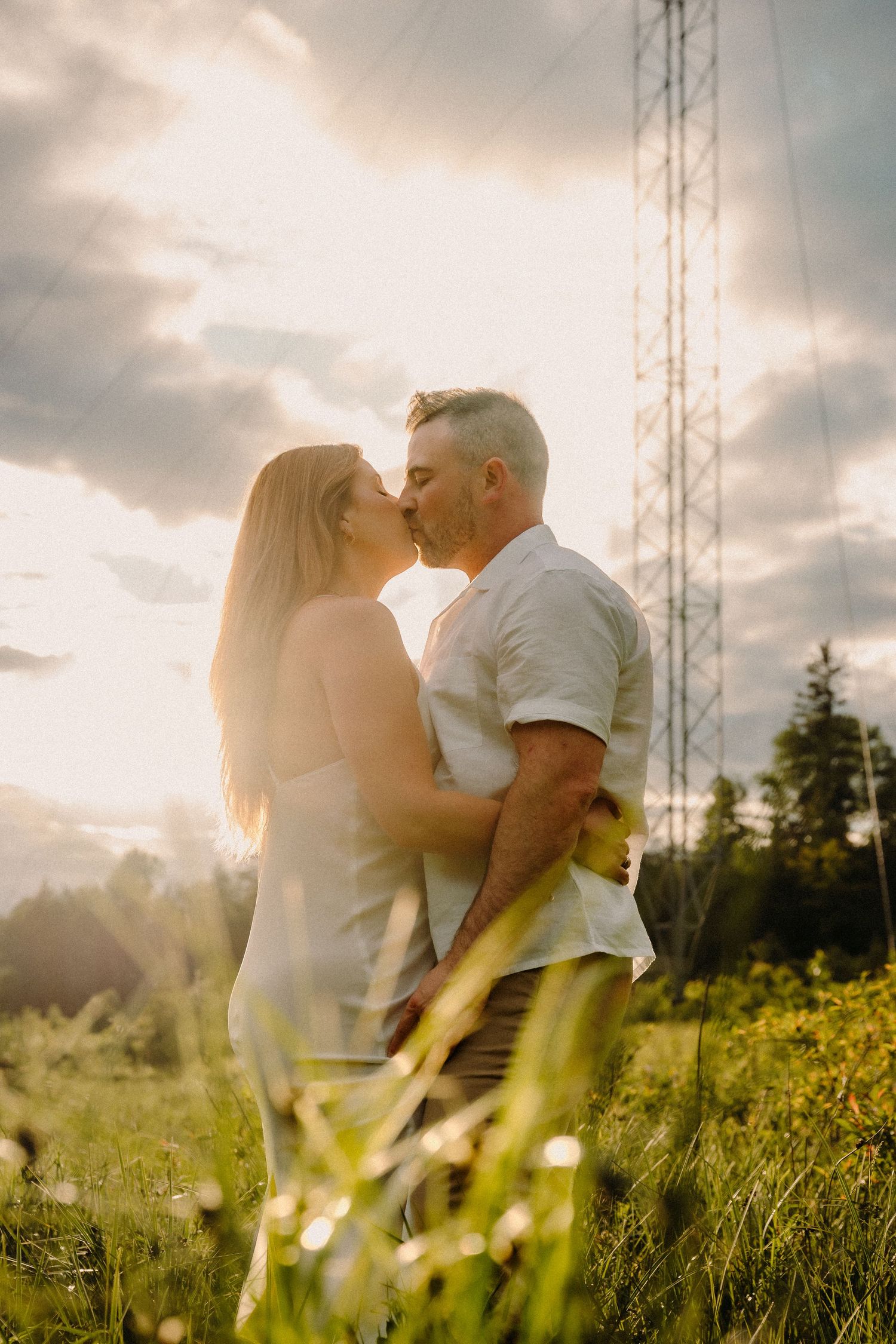 Couple sharing a romantic kiss in a sunlit meadow with dramatic cloudy sky and transmission tower backdrop.