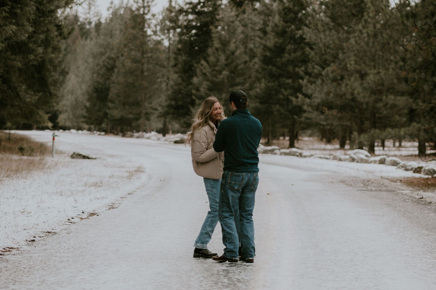 Couple embracing on a snowy road surrounded by pine trees in a winter forest landscape.