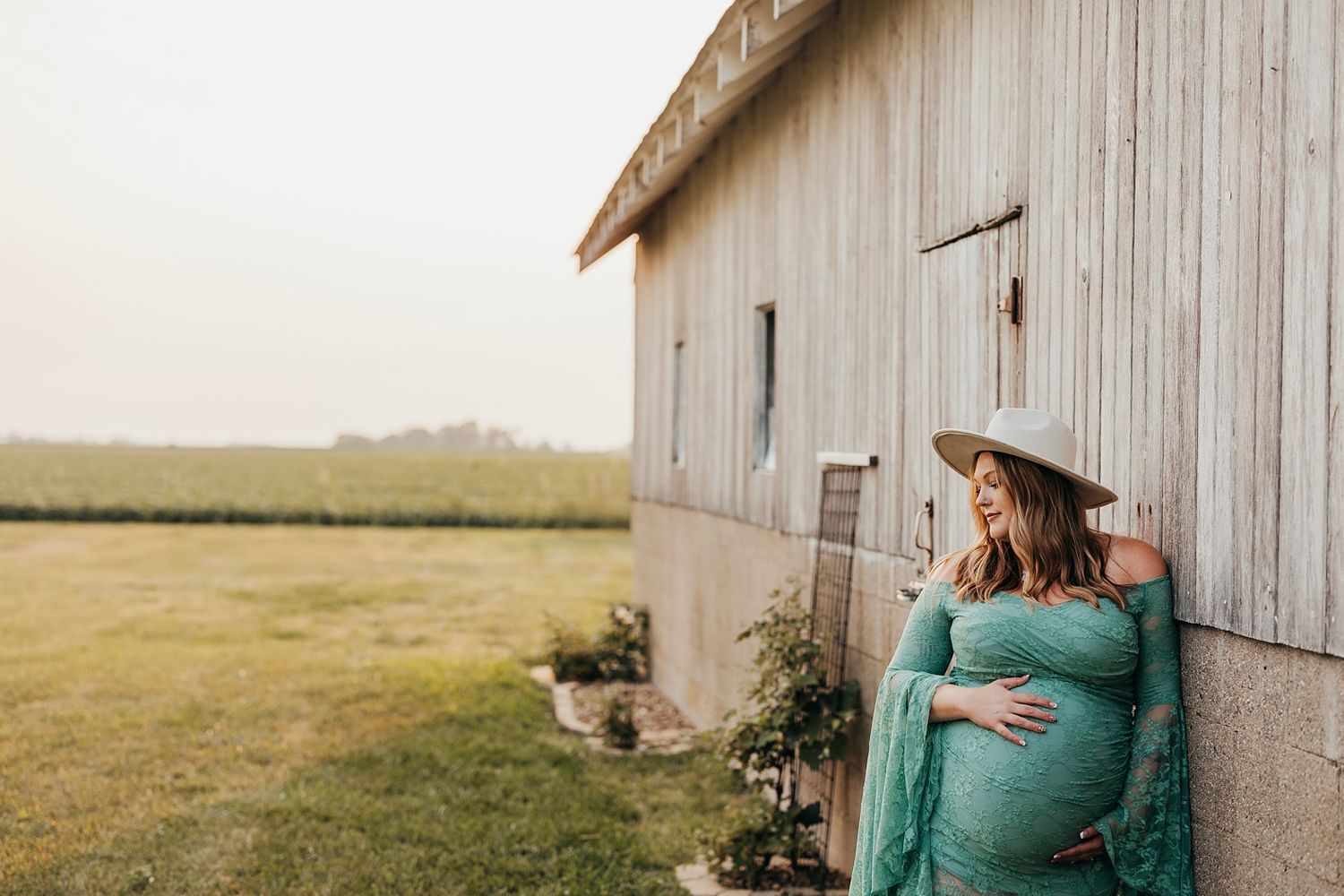 Pregnant woman in teal lace gown and hat leaning against rustic barn wall with green fields in background.
