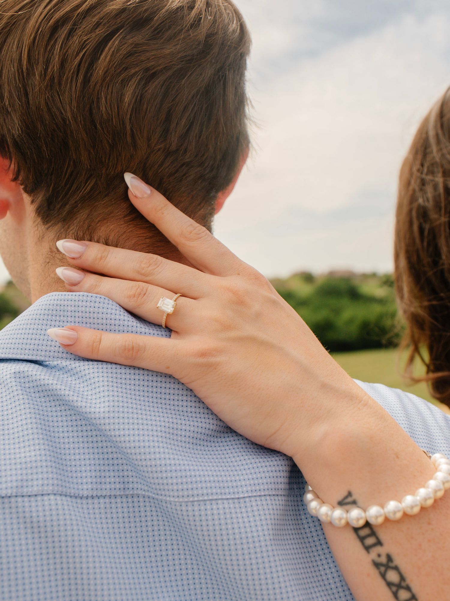Couple embracing outdoors, woman showing diamond engagement ring and pearl bracelet with Roman numeral tattoo on wrist.