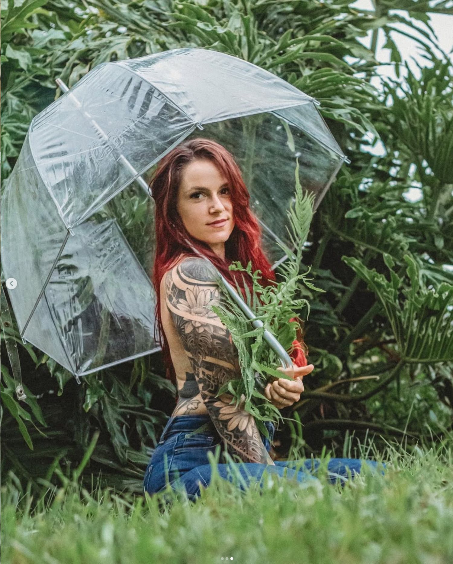 Red-haired tattooed woman holding a clear bubble umbrella outdoors among lush green tropical plants.