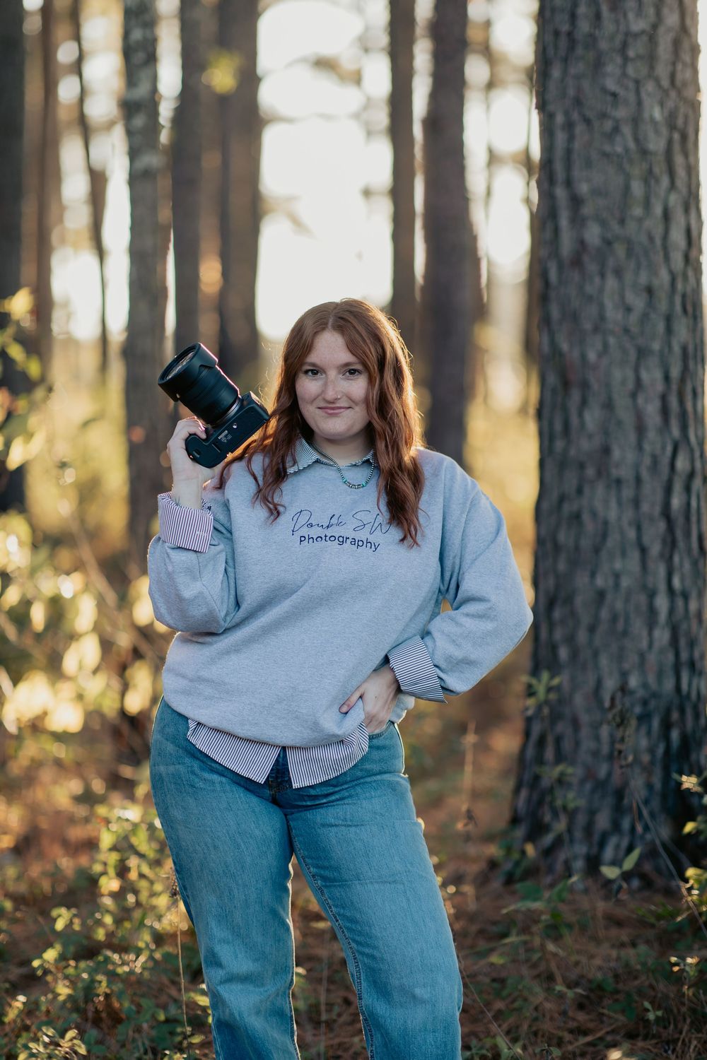 Red-haired female photographer holding camera in forest, wearing gray photography sweatshirt and jeans.