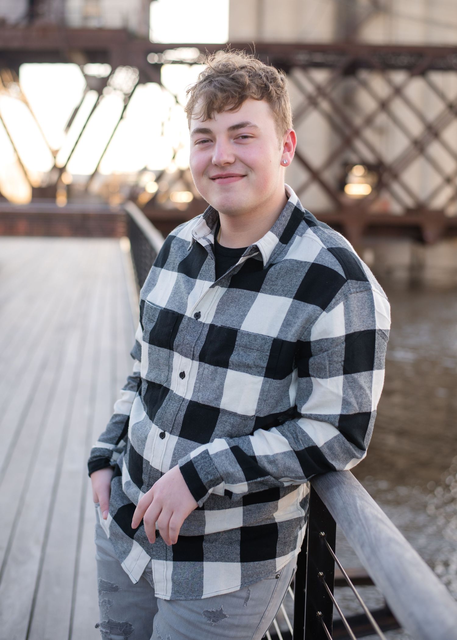 Smiling teen in black and white plaid shirt poses on wooden boardwalk near rustic metal bridge at sunset.