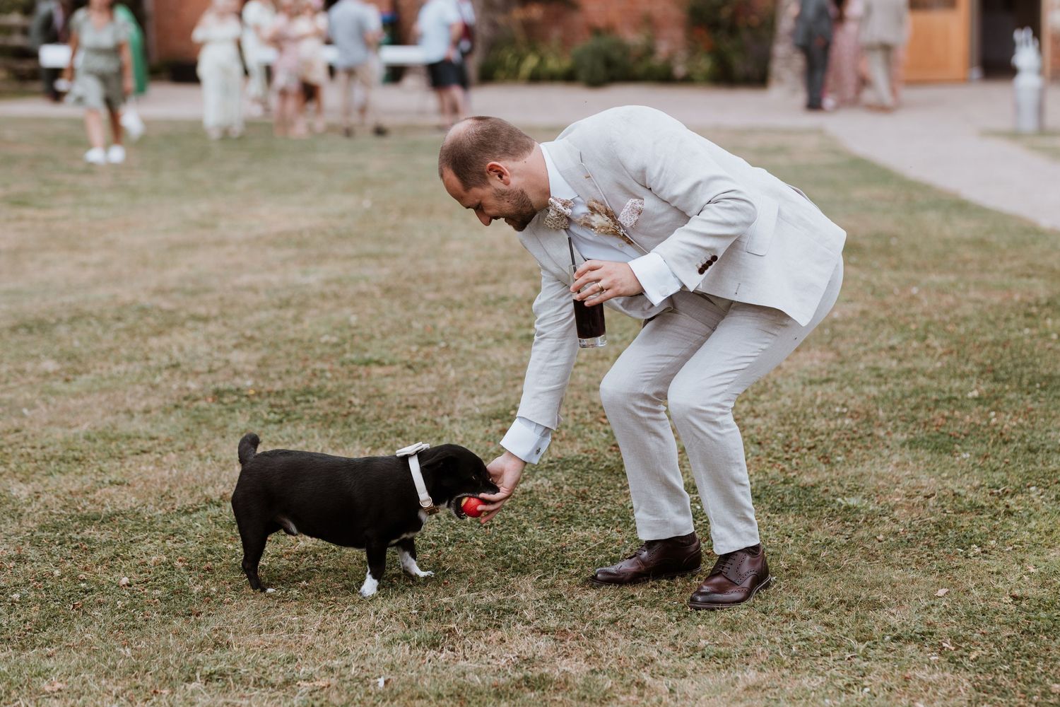 Groom in light gray suit bending down to interact with a small black pug dog on a grass lawn at an outdoor wedding.