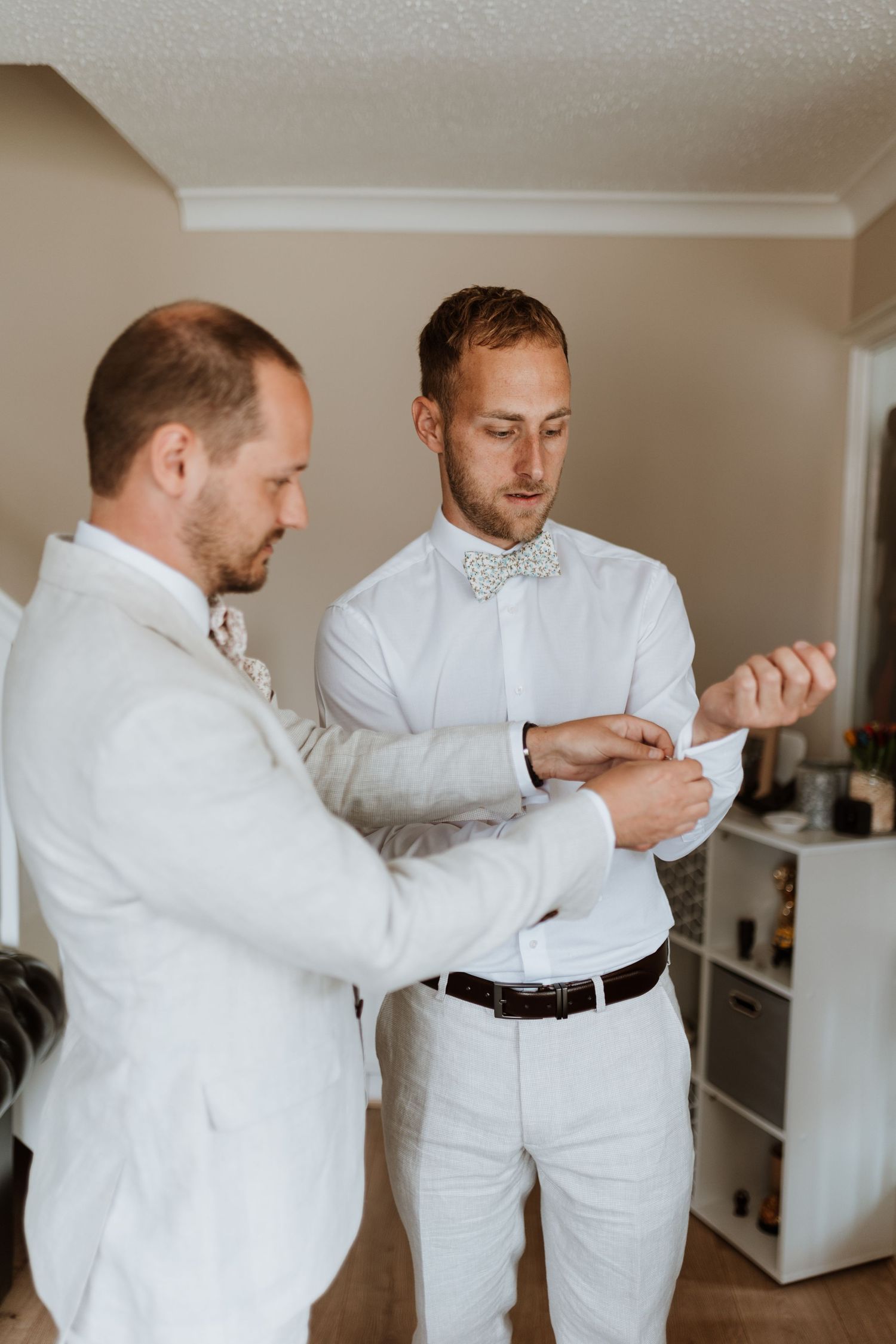 Two groomsmen in white suits help each other with cufflinks while preparing for a wedding ceremony