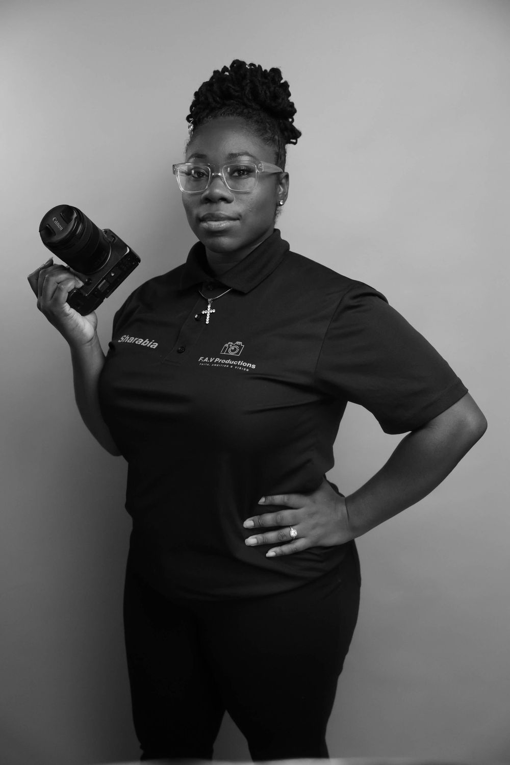 Black and white portrait of a female photographer holding a camera, wearing a black polo shirt with a cross necklace.