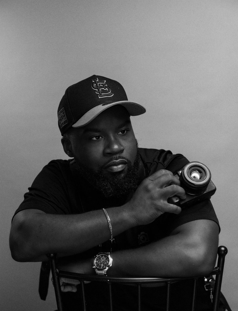 Black and white portrait of a bearded man in a cap holding a camera, seated on a chair in a studio setting.