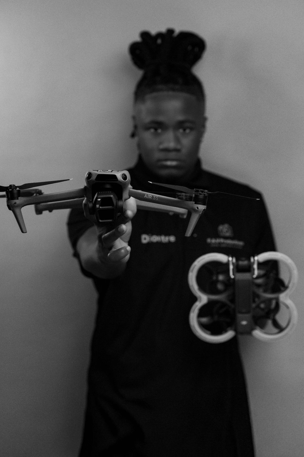 Black and white photo of a young man holding two drones toward the camera in a studio setting.