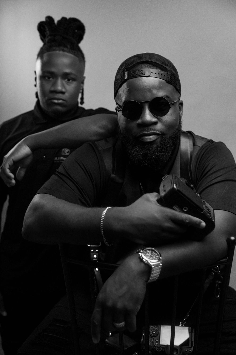 Black and white portrait of two men in dark clothing, one wearing sunglasses and a watch, posing confidently.