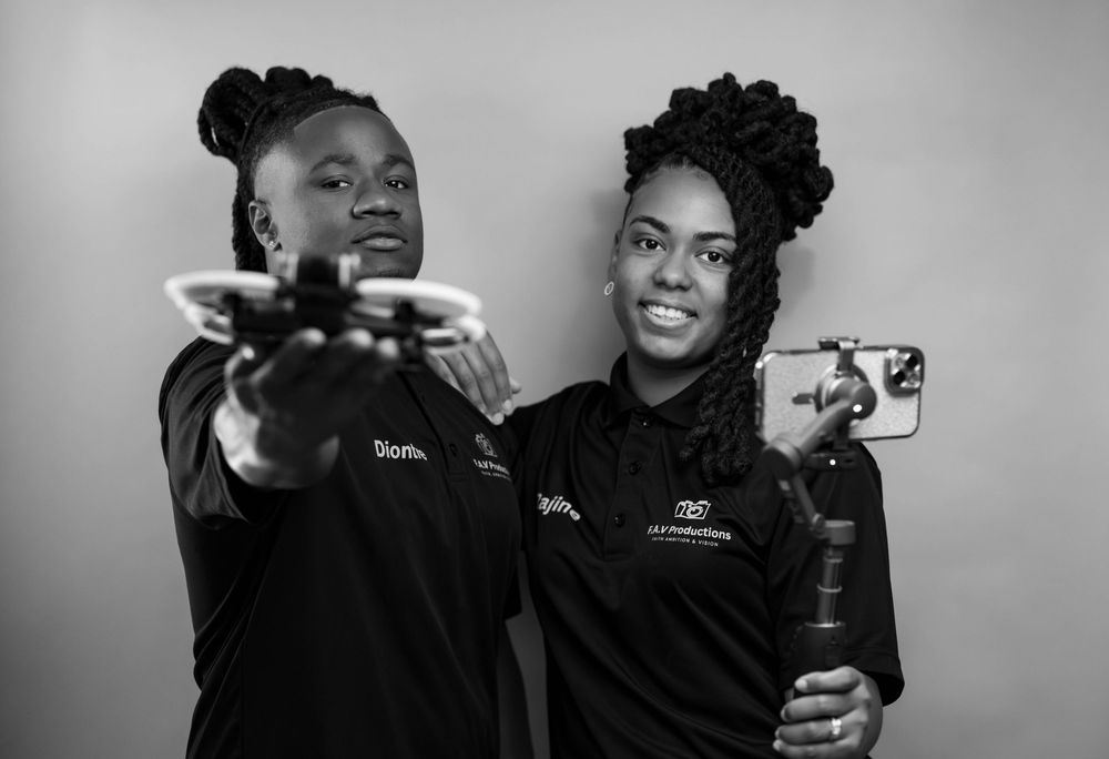 Two young women in black shirts hold a drone and smartphone, posing confidently in a black and white studio photo.