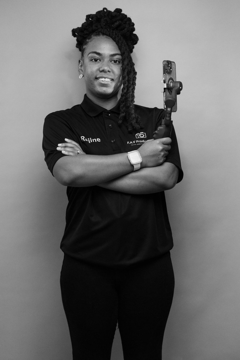 Black and white portrait of a smiling woman in branded polo shirt holding a camera, arms crossed confidently.
