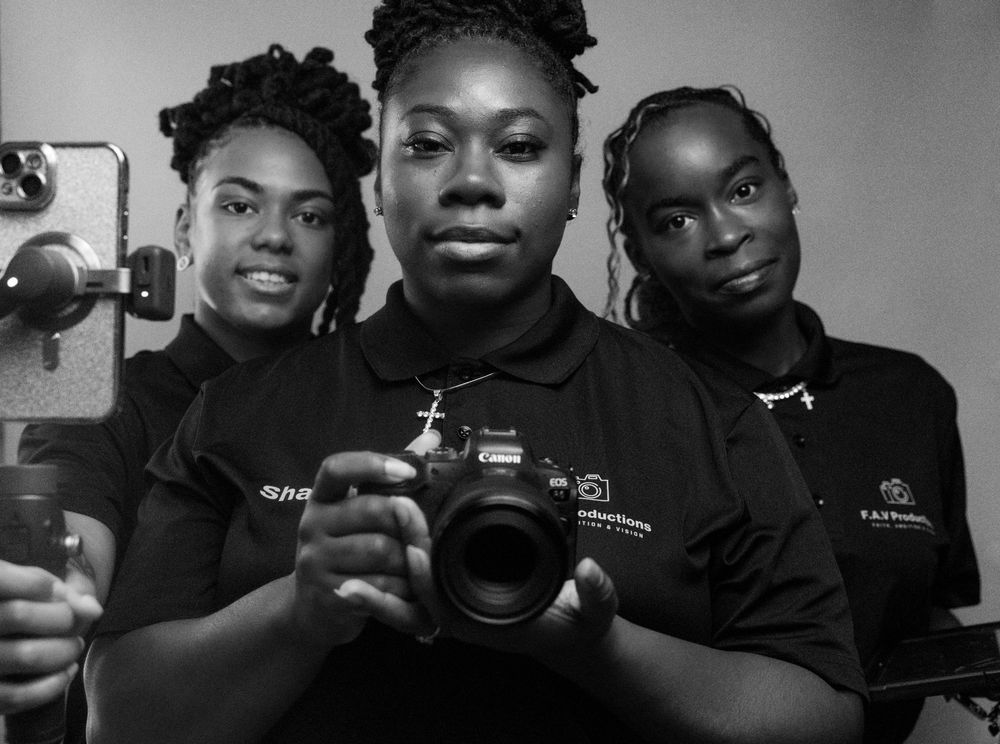 Three young women in matching black polo shirts posing with a Canon camera in a black and white photo.