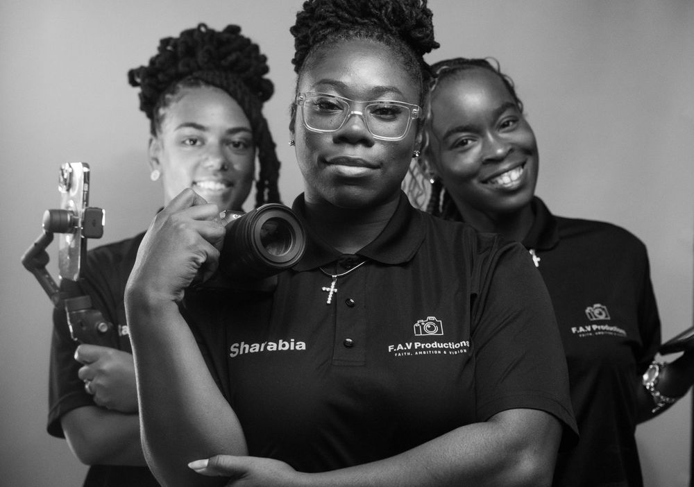 Three women in matching black polo shirts pose together holding photography equipment in a black and white photo.