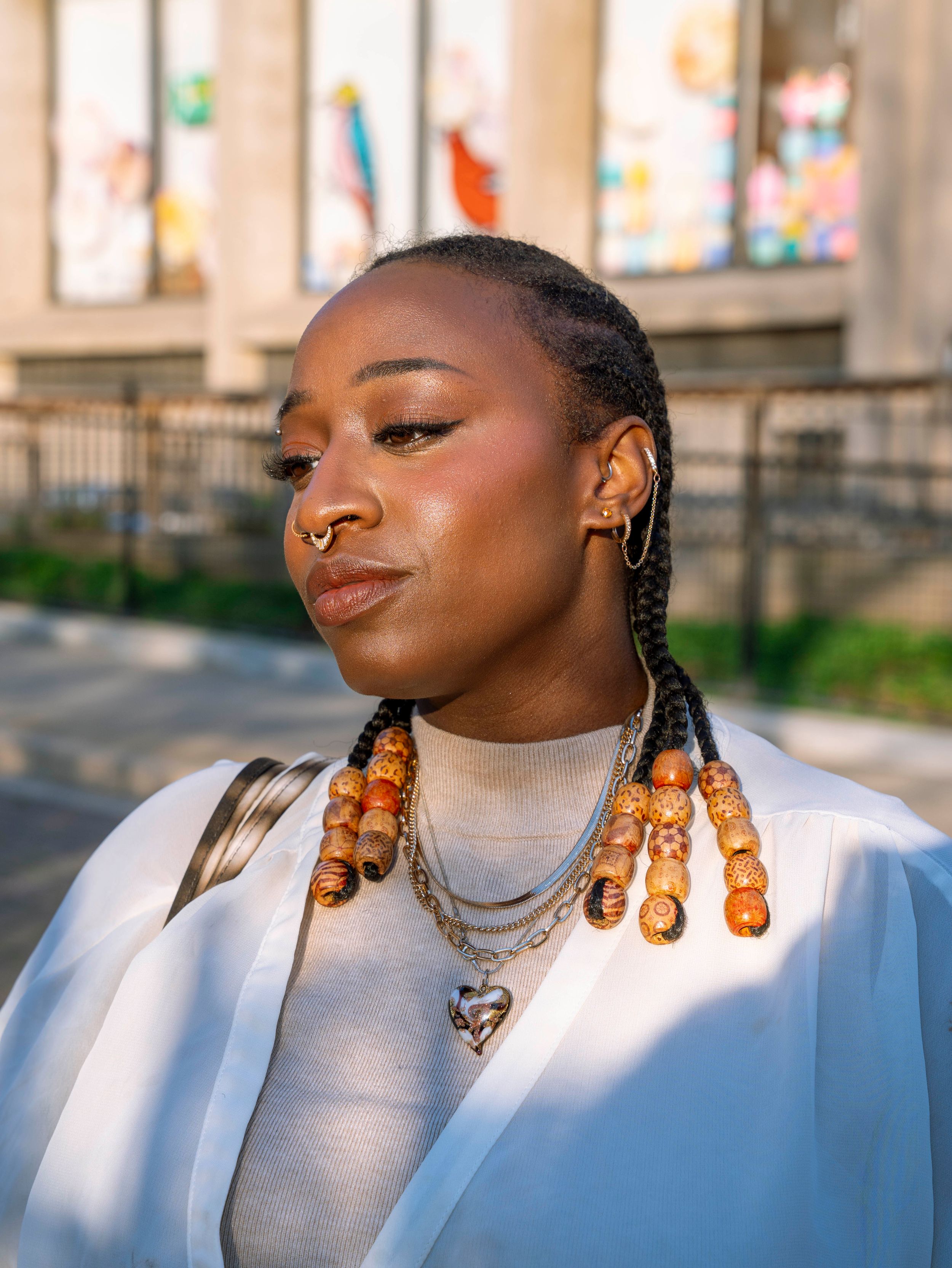 Close-up of Black woman with beaded braids, nose ring, hoop earrings, and layered gold necklaces outdoors.