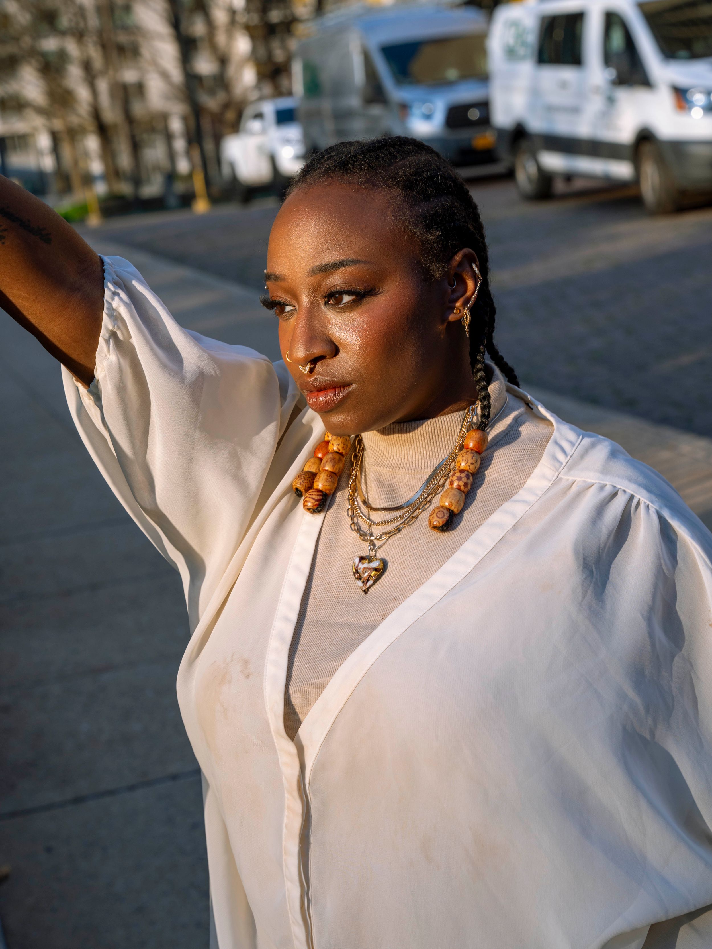 Black woman in white linen shirt with beaded braids and layered necklaces poses on sunny urban street.