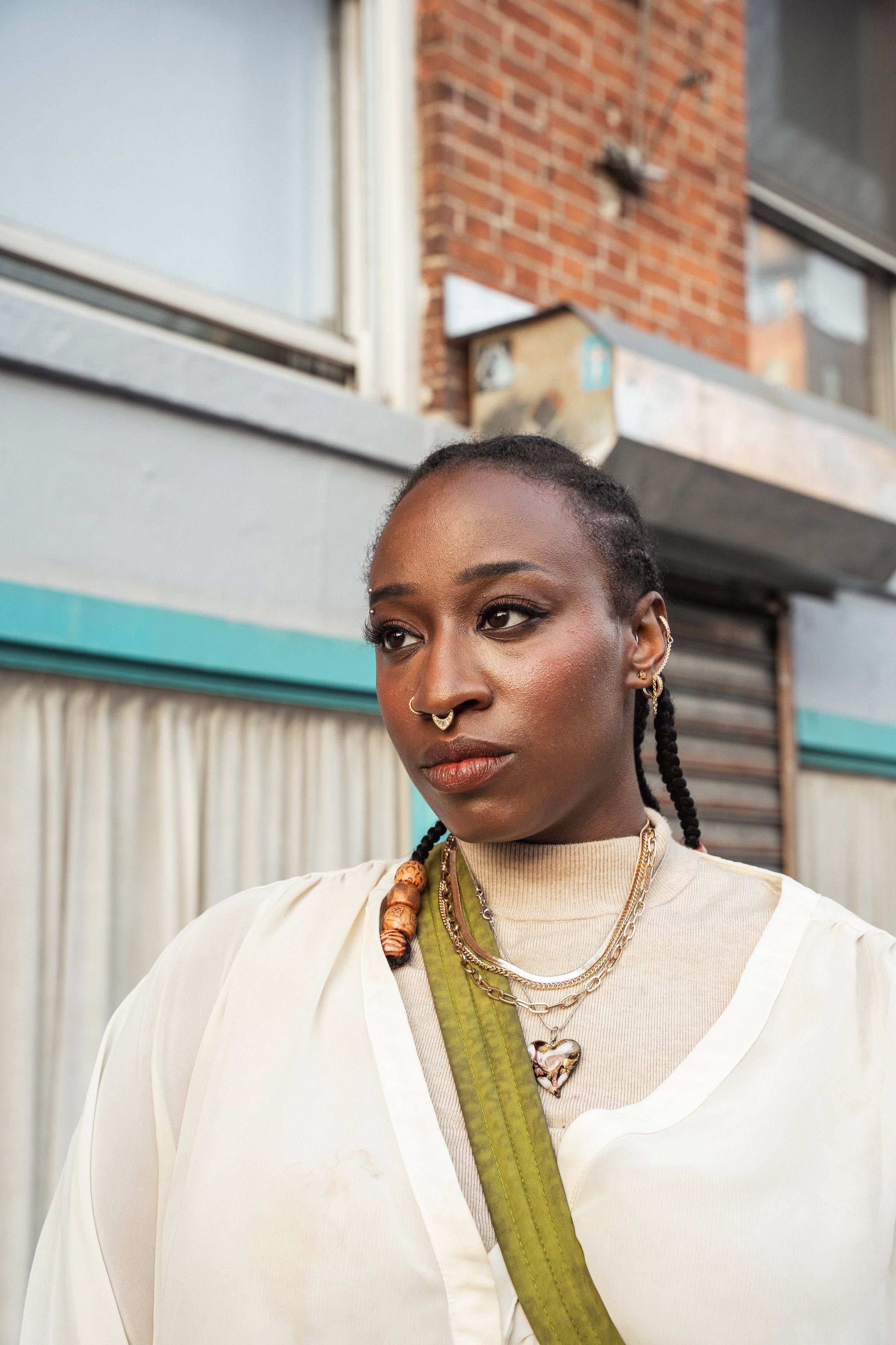 Young Black woman with braids, nose ring, and layered gold necklaces poses outdoors in white top.