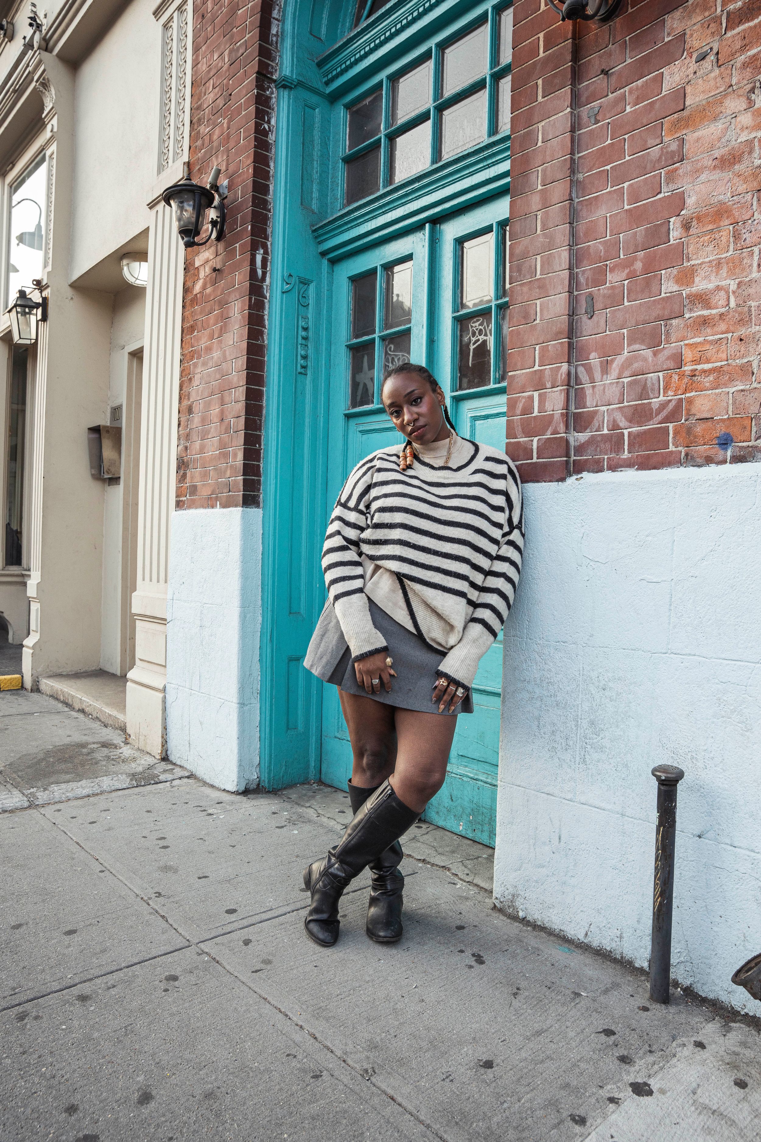Black woman in striped oversized sweater, gray skirt, and tall boots stands by teal door on sidewalk.