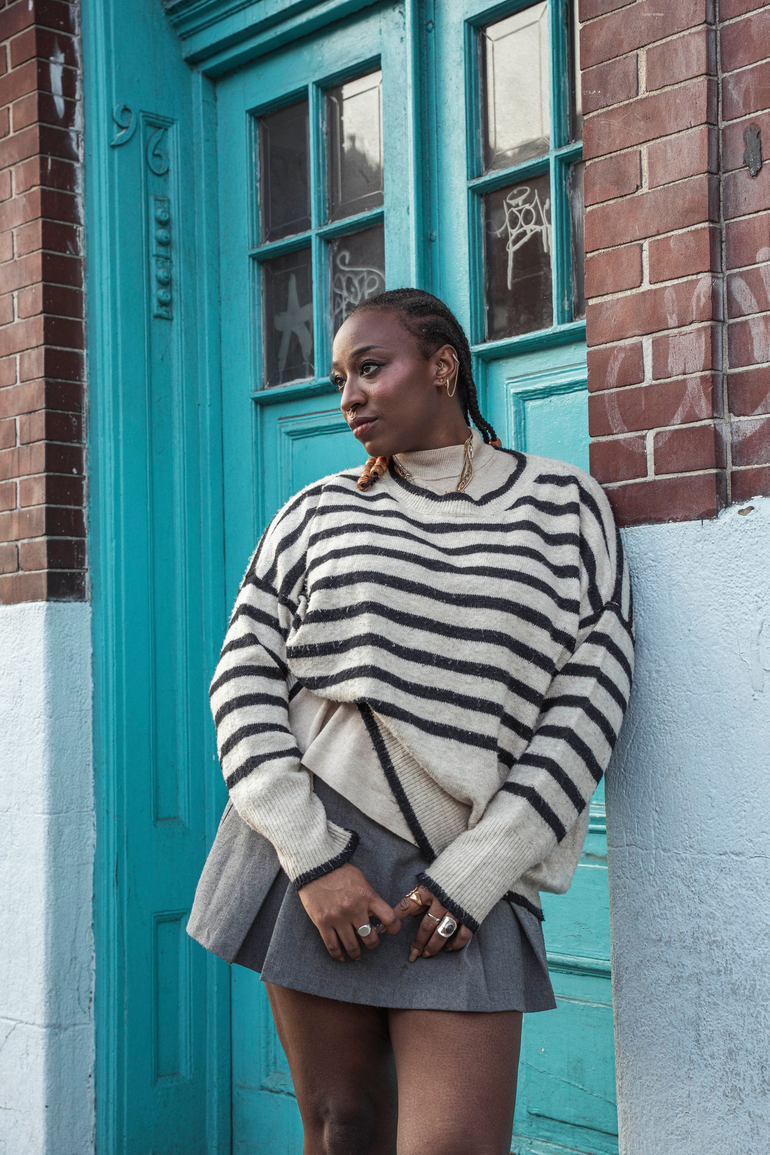 Black woman in oversized striped sweater and gray mini skirt leans against teal door on brick wall.