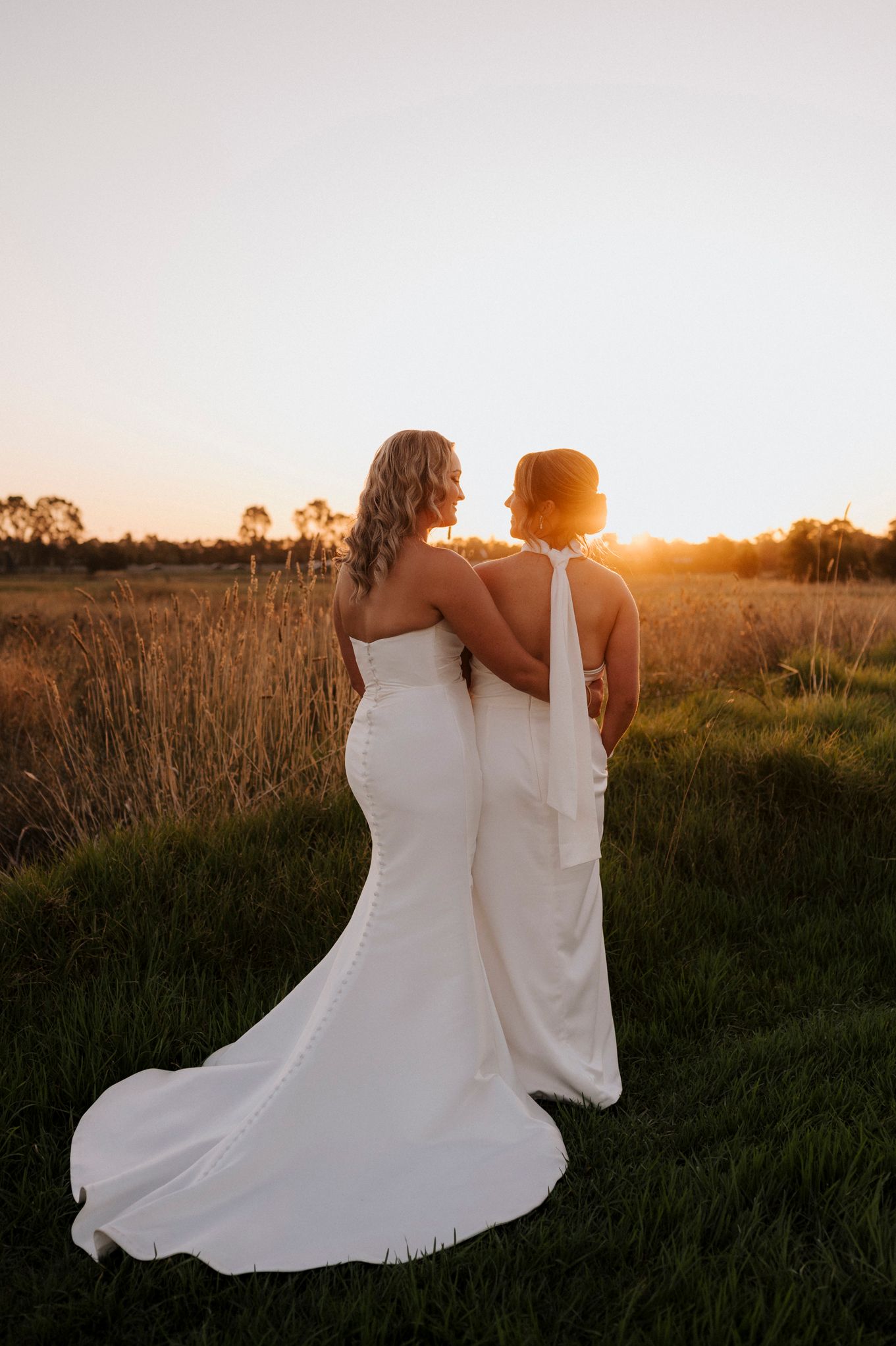 Two brides in white gowns embrace at sunset in a golden field, sharing a romantic wedding moment.