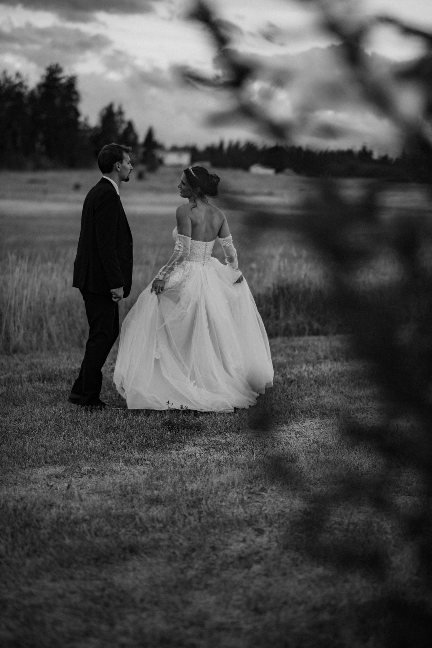 Black and white photo of bride and groom holding hands in a field at dusk, bride in elegant off-shoulder gown.