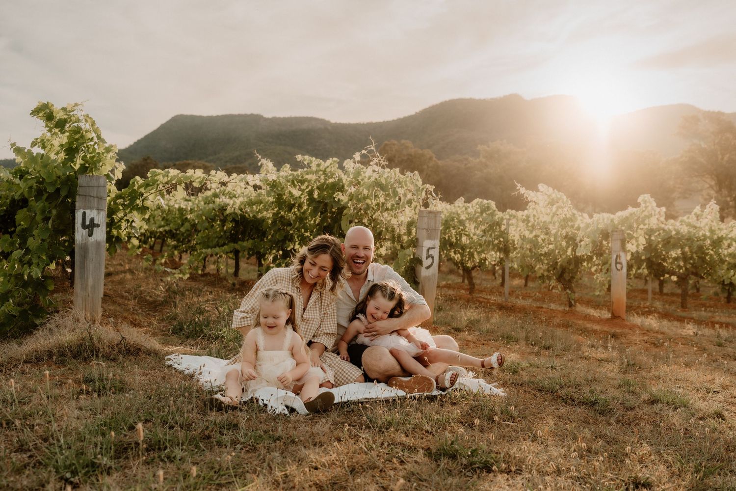 Happy family of four sitting on a blanket in a vineyard at golden hour with mountain backdrop.