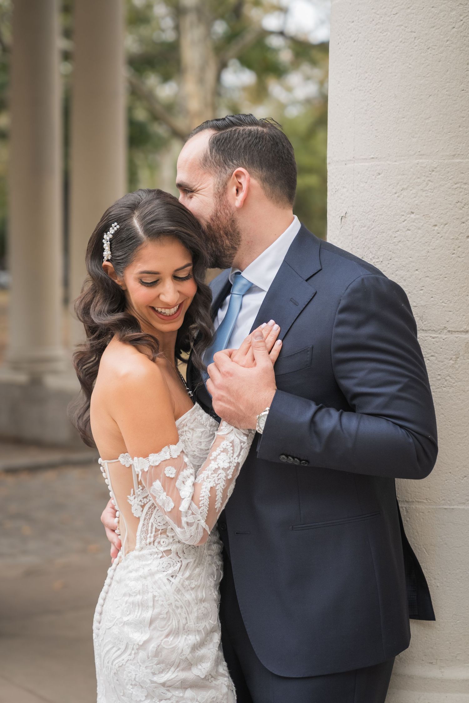 Bride in lace off-shoulder gown laughs as groom in navy suit kisses her cheek near elegant stone columns.