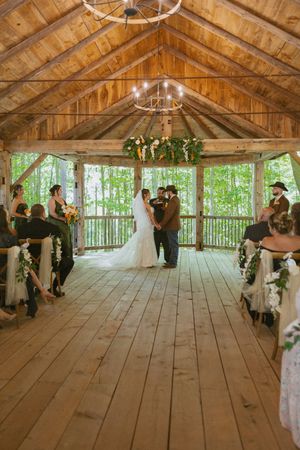 Rustic barn wedding ceremony with couple at floral arch, wood plank floors, chandelier, and guests seated in forest pavilion.