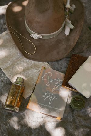 Rustic wedding flat lay with cowboy hat, save the date card, perfume bottle, and ring on stone surface.