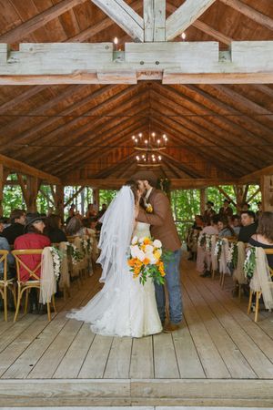 Bride and groom share first kiss at rustic barn wedding ceremony with orange floral bouquet.