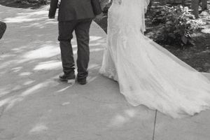 Black and white photo of a bride in a white gown and groom in a suit walking together on a paved path.