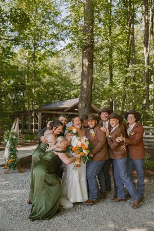 Wedding party in forest setting, bridesmaids in green satin, groomsmen in brown suits and cowboy hats, bride holding orange bouquet.