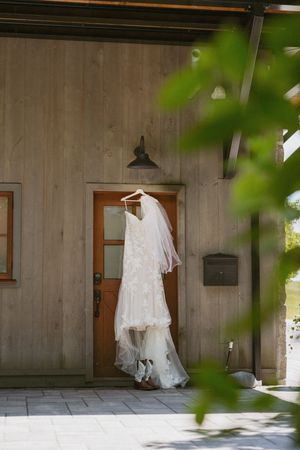 White lace wedding dress with veil hanging on a wooden barn door with cowboy boots below.