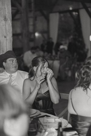 Emotional wedding guest wiping tears during reception speech inside rustic barn venue, black and white photo.