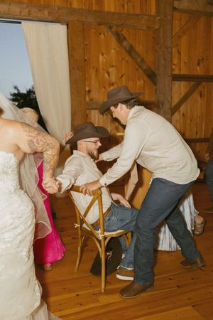 Two men in cowboy hats share a lively moment at a rustic barn wedding reception on a wooden dance floor.