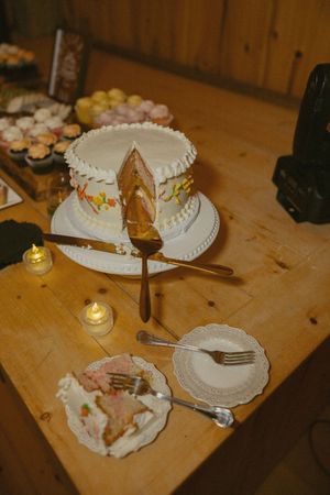 White frosted wedding cake with a slice removed, served on a wooden table with cupcakes and lit candles nearby.