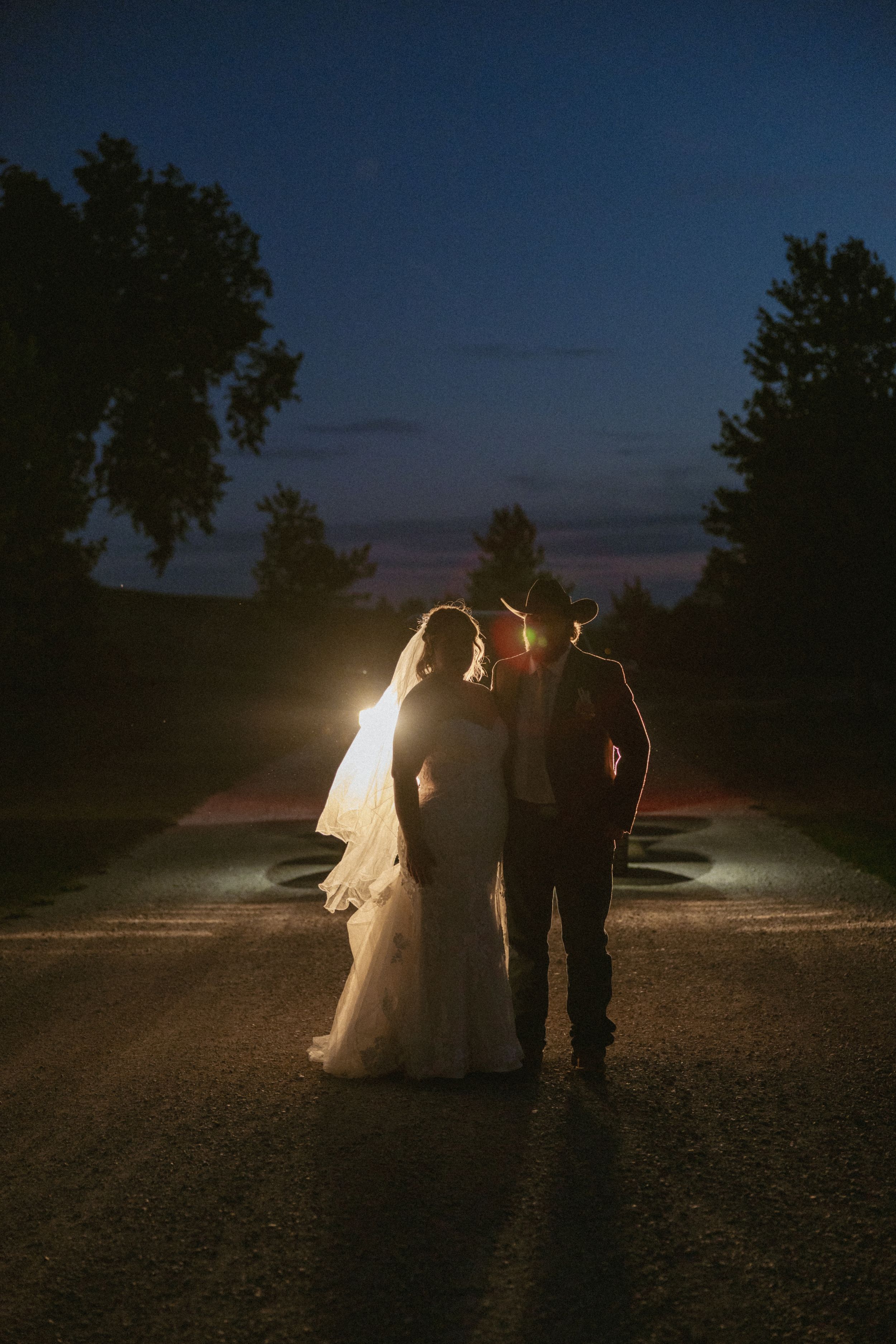 Bride and groom silhouetted against car headlights at dusk, surrounded by trees in a romantic night wedding portrait.