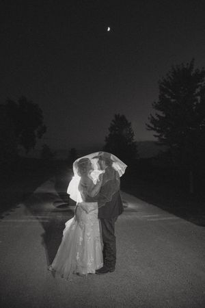 Black and white photo of a bride and groom embracing under a veil at night with a crescent moon above.