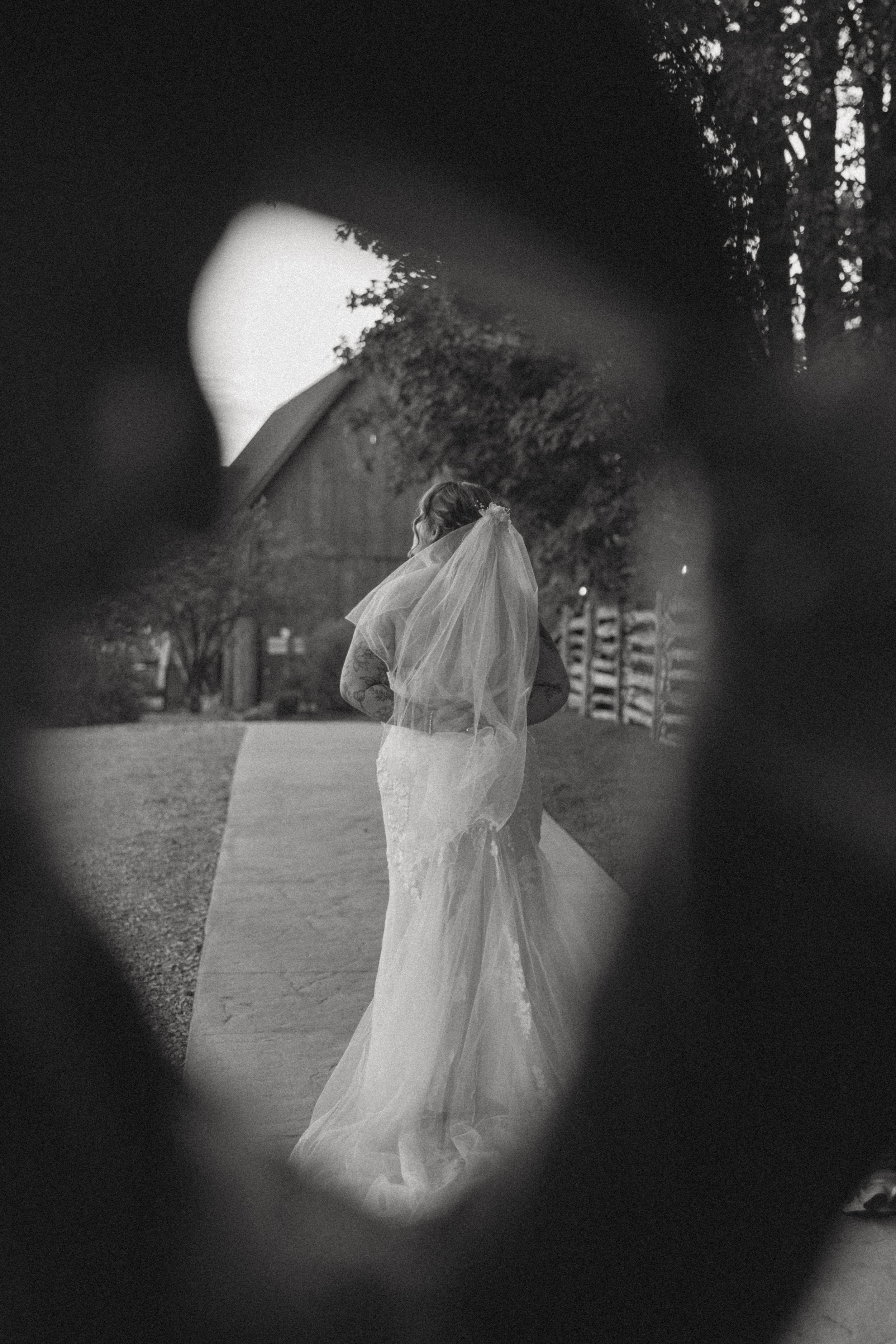 Black and white photo of a bride in a lace gown and veil, framed through a dark foreground, standing near a rustic barn.