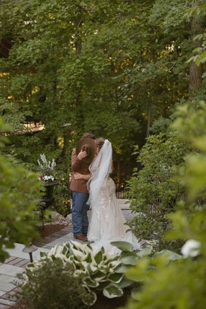 Bride and groom embrace in a lush garden pathway surrounded by green trees and plants during their outdoor wedding ceremony.