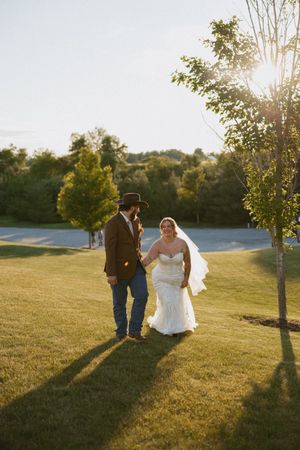 Bride and groom walk hand-in-hand on lush green lawn at golden hour, groom in cowboy hat and brown jacket.