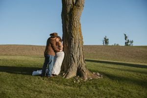 Couple sharing a romantic kiss behind a large tree trunk in a green field under a clear blue sky.