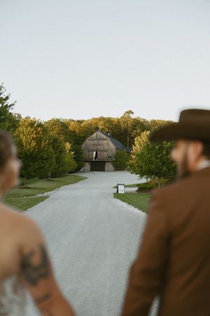 Bride and groom in cowboy hat walking toward rustic round barn venue along gravel path at golden hour.