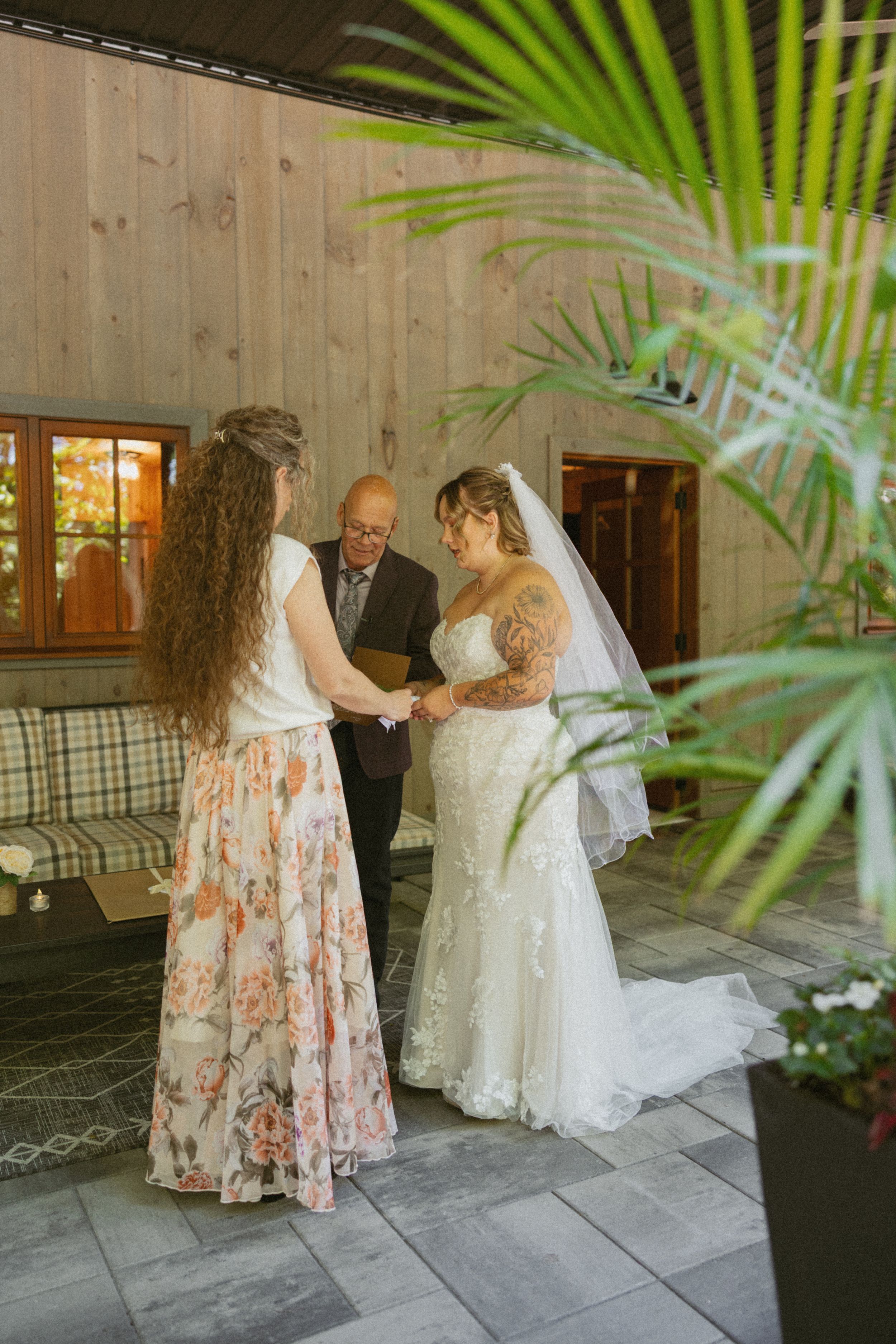 Two brides exchange vows with an officiant during an intimate indoor wedding ceremony near tropical plants.