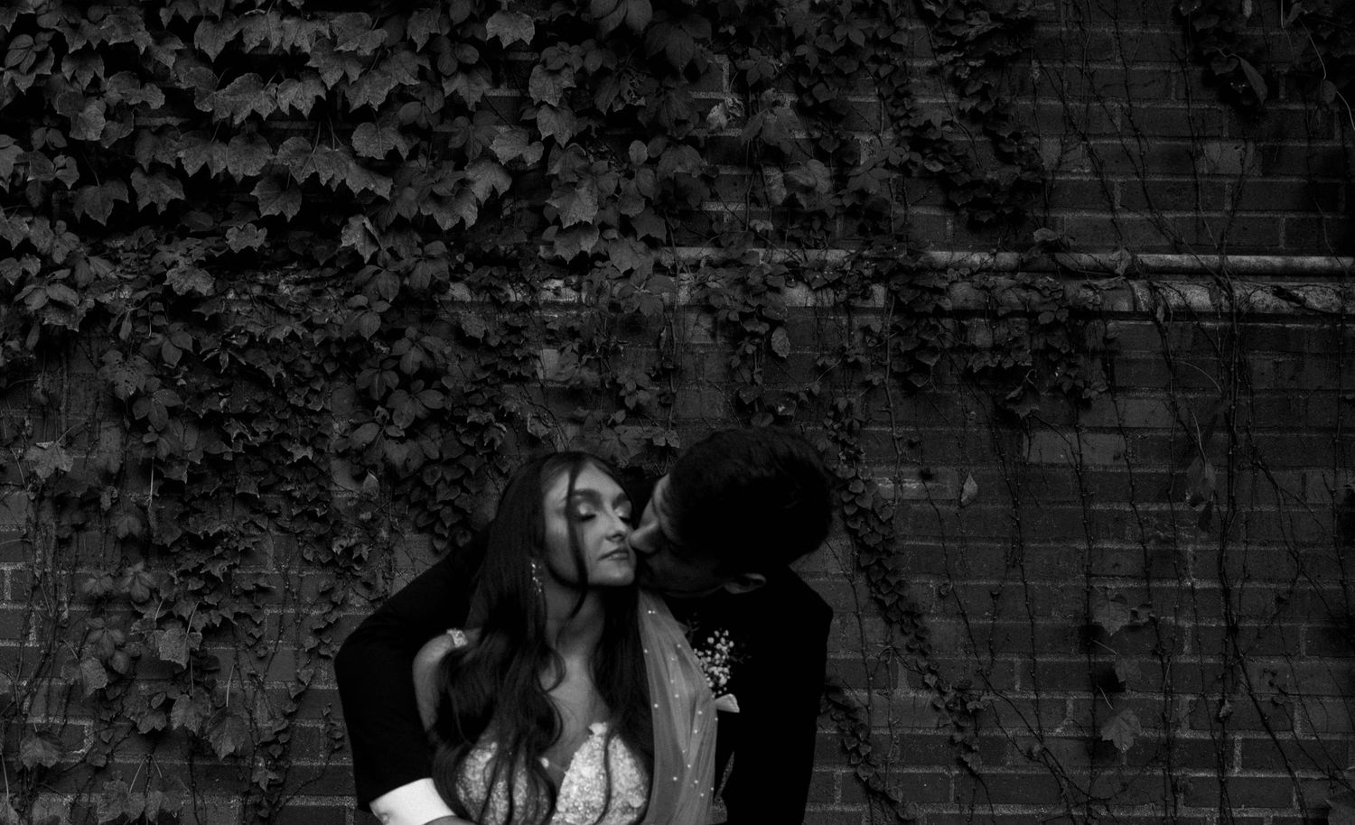 Black and white photo of a couple kissing against an ivy-covered brick wall.