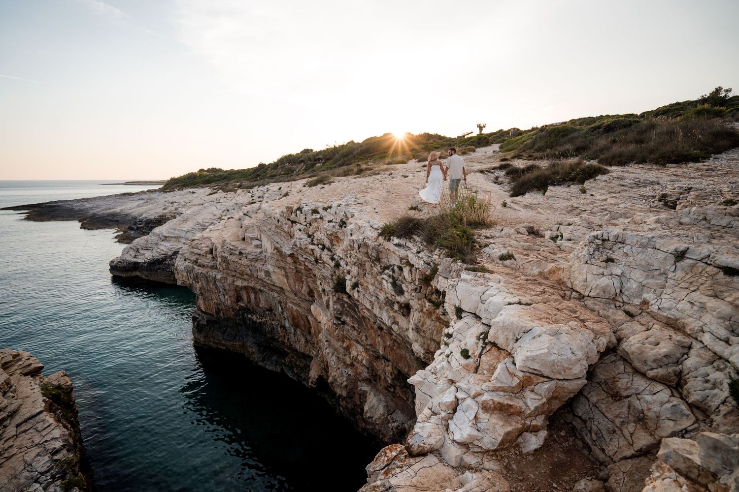 Couple in white attire standing on rocky coastal cliffs at sunset with calm sea waters below.
