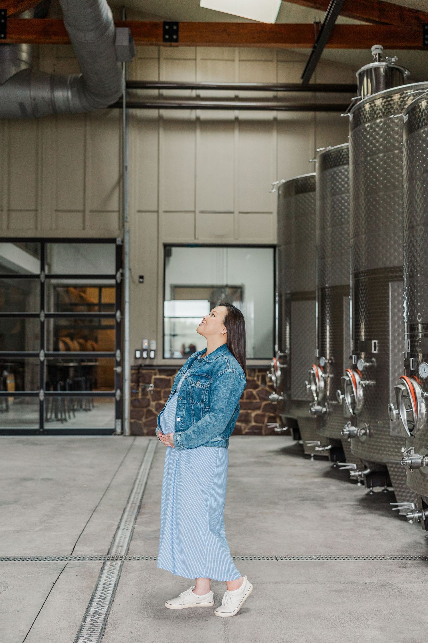 Pregnant woman in denim jacket and blue skirt stands in modern winery beside stainless steel fermentation tanks.